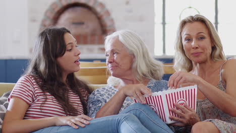 Mother-With-Adult-Daughter-And-Teenage-Granddaughter-Eating-Popcorn-Watching-Movie-On-Sofa-At-Home