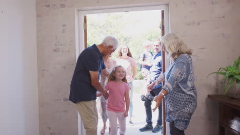 Grandparents-Open-Front-Door-Of-House-To-Welcome-Multi-Generation-Family-On-Visit