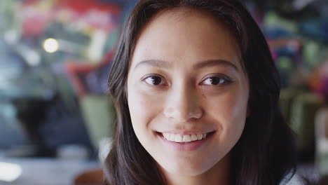 Young-female-Asian-creative-smiling-to-camera-in-front-of-mural-in-workplace,-head-shot,-close-up