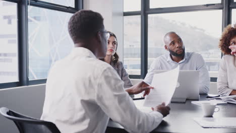 Business-team-at-a-table-in-meeting-room-discussing-document,-middle-aged-white-businessman-standing
