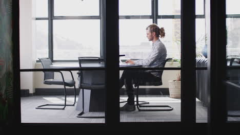Young-white-businessman-with-ponytail-working-alone-in-an-office,-seen-through-glass-wall,-zoom-in