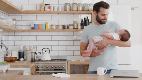 Multi-Tasking-Father-Holds-Sleeping-Baby-Son-Whilst-Cleaning-And-Working-On-Laptop-In-Kitchen