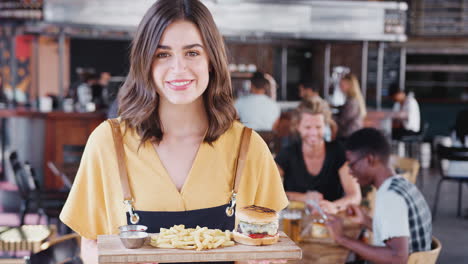 Portrait-Of-Waitress-Serving-Food-In-Busy-Bar-Restaurant-Smiling-At-Camera