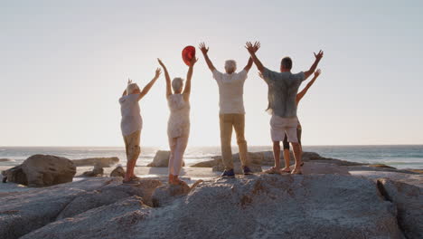 Rear-View-Of-Senior-Friends-Standing-On-Rocks-And-Raising-Arms-On-Group-Vacation-Looking-Out-To-Sea