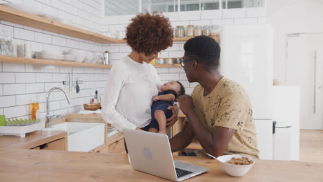 Busy-Family-In-Kitchen-At-Breakfast-With-Father-Working-On-Laptop-And-Mother-Caring-For-Baby-Son