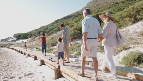 Familia-Multigeneracional-En-Vacaciones-De-Verano-Caminando-Por-Un-Paseo-Marítimo-De-Madera-Camino-A-La-Playa