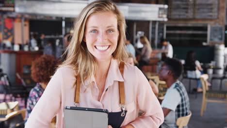 Portrait-Of-Waitress-Holding-Digital-Tablet-In-Busy-Bar-Restaurant-Smiling-At-Camera