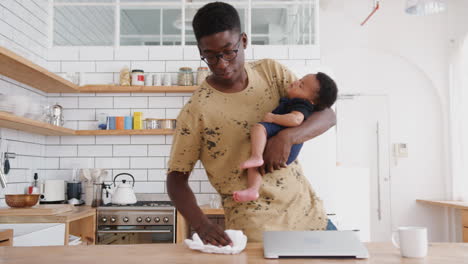 Multi-Tasking-Father-Holds-Sleeping-Baby-Son-Whilst-Cleaning-And-Working-On-Laptop-In-Kitchen