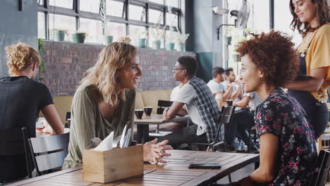 Two-Female-Friends-Sitting-At-Table-In-Coffee-Shop-Being-Served-By-Waitress