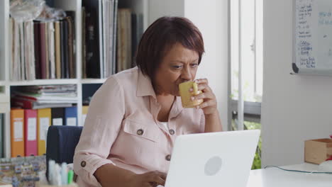 Mature-Woman-With-Laptop-Working-In-Home-Office-Drinking-Coffee