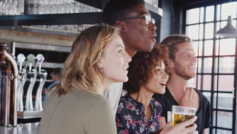 Group-Of-Male-And-Female-Friends-Celebrating-Whilst-Watching-Game-On-Screen-In-Sports-Bar