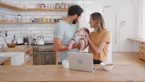 Busy-Family-In-Kitchen-At-Breakfast-With-Mother-Working-On-Laptop-And-Father-Caring-For-Baby-Son