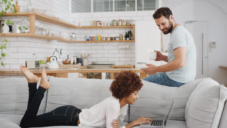Man-With-Hot-Drink-Talks-To-Woman-Relaxing-Lying-On-Sofa-At-Home-Looking-At-Laptop