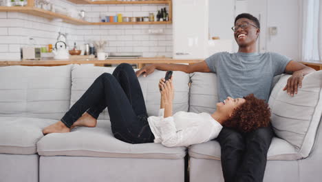 Young-Couple-Relaxing-On-Sofa-At-Home-Looking-At-Mobile-Phone-And-Laughing-Together