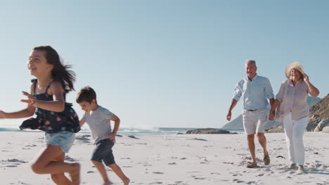 Grandparents-With-Grandchildren-Walking-Along-Sandy-Beach-On-Summer-Vacation