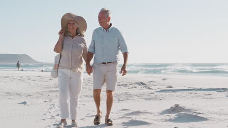Senior-Couple-Walking-Along-Beautiful-Sandy-Beach-On-Summer-Vacation
