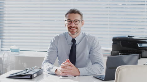Portrait-Of-Mature-Male-Doctor-Sitting-Behind-Desk-In-Office