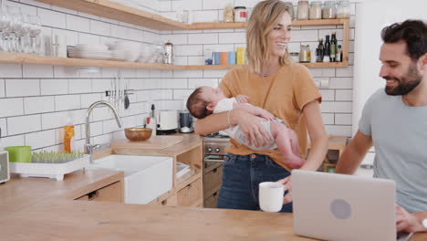 Multitasking-Mutter-Hält-Ihren-Kleinen-Sohn-Und-Kocht-Heiße-Getränke,-Während-Der-Vater-Seinen-Laptop-Benutzt-Und-Frühstückt