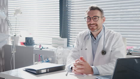 Portrait-Of-Smiling-Mature-Male-Doctor-In-White-Coat-With-Stethoscope-Sitting-Behind-Desk-In-Office