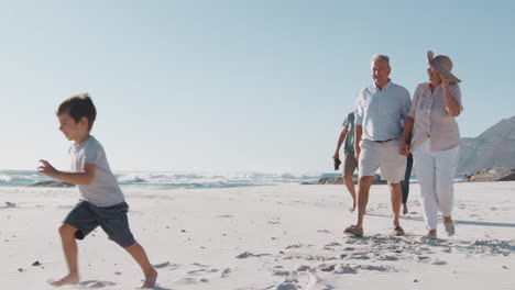 Multi-Generation-Family-Walking-Along-Sandy-Beach-On-Summer-Vacation