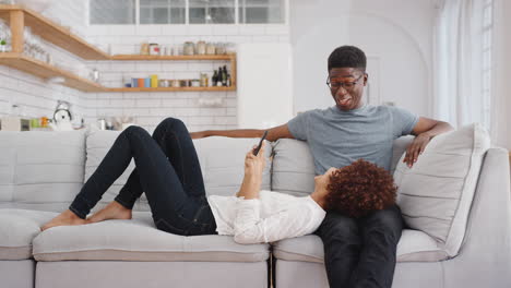 Young-Couple-Relaxing-On-Sofa-At-Home-Looking-At-Mobile-Phone-And-Laughing-Together