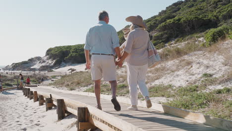 Älteres-Paar-Im-Sommerurlaub,-Das-Auf-Dem-Weg-Zum-Strand-Entlang-Der-Holzpromenade-Spaziert