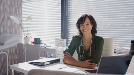 Portrait-Of-Smiling-Female-Doctor-With-Stethoscope-Sitting-Behind-Desk-In-Office