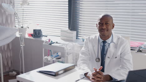 Portrait-Of-Male-Doctor-Wearing-White-Coat-With-Stethoscope-Sitting-At-Desk-In-Office