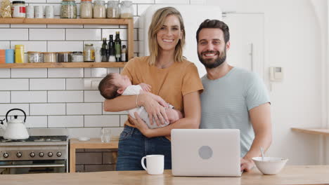 Portrait-Of-Family-In-Kitchen-At-Breakfast-With-Mother-Caring-For-Baby-Son-And-Father-Using-Laptop