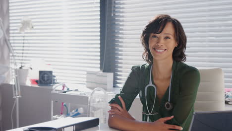 Portrait-Of-Smiling-Female-Doctor-With-Stethoscope-Sitting-Behind-Desk-In-Office