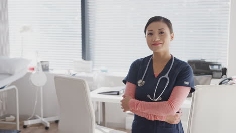 Portrait-Of-Smiling-Female-Doctor-Wearing-Scrubs-With-Stethoscope-Standing-By-Desk-In-Office