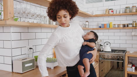 Multi-Tasking-Mother-Holds-Sleeping-Baby-Son-Whilst-Cleaning-And-Working-On-Laptop-In-Kitchen