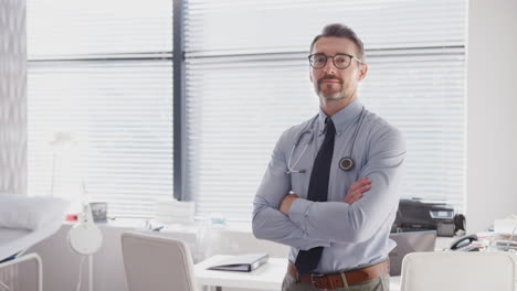 Portrait-Of-Smiling-Mature-Male-Doctor-Standing-By-Desk-In-Office