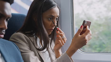 Businesswoman-Sitting-In-Train-Commuting-To-Work-Putting-On-Make-Up-Using-Mobile-Phone-As-Mirror