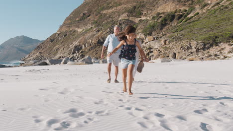 Grandparents-With-Granddaughter-Walking-Along-Sandy-Beach-On-Summer-Vacation