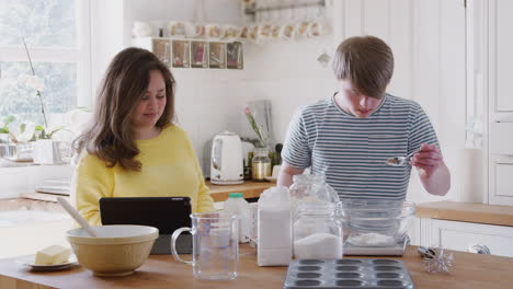 Young-Downs-Syndrome-Couple-Following-Recipe-On-Digital-Tablet-To-Bake-Cake-In-Kitchen-At-Home
