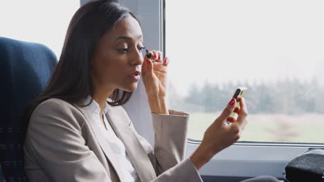 Mujer-De-Negocios-Sentada-En-El-Tren-Yendo-Al-Trabajo-Maquillándose-Usando-El-Teléfono-Móvil-Como-Espejo