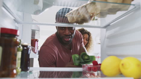 Blick-Aus-Dem-Inneren-Des-Kühlschranks,-Während-Ein-Paar-Die-Tür-öffnet-Und-Die-Einkaufstüte-Mit-Lebensmitteln-Auspackt