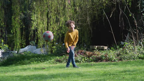 Young-Boy-Kicking-Soccer-Ball-In-Garden-At-Home