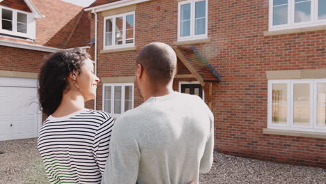Rear-View-Of-Loving-Couple-Standing-Outside-New-Home-On-Moving-Day-Looking-At-House