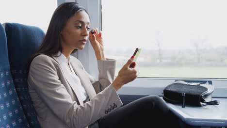Mujer-De-Negocios-Sentada-En-El-Tren-Yendo-Al-Trabajo-Maquillándose-Usando-El-Teléfono-Móvil-Como-Espejo