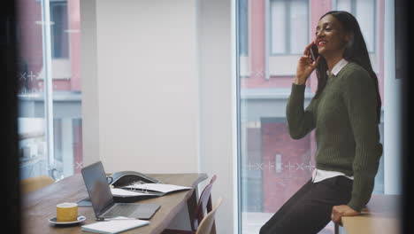 Businesswoman-Sitting-On-Desk-In-Meeting-Room-Talking-On-Mobile-Phone
