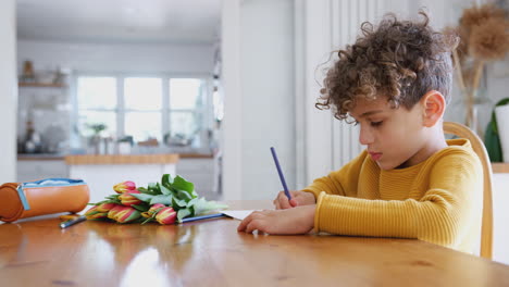 Kleiner-Junge-Zu-Hause-Mit-Blumenstrauß,-Der-In-Eine-Muttertagskarte-Schreibt