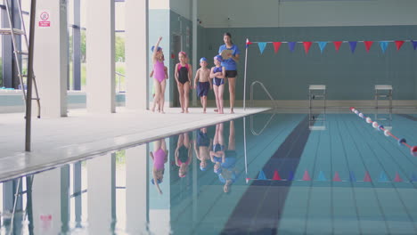 Female-Coach-Walks-Children-In-Swimming-Class-Along-Edge-Of-Indoor-Pool