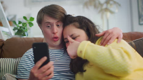 Junges-Paar-Mit-Down-Syndrom-Sitzt-Auf-Dem-Sofa-Und-Macht-Mit-Dem-Handy-Zu-Hause-Ein-Selfie
