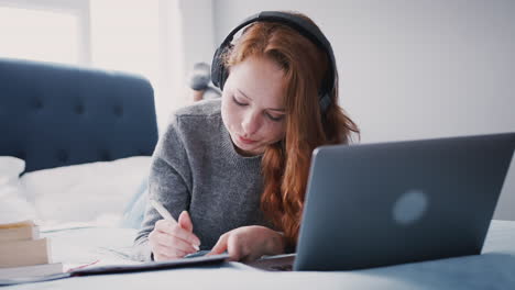 Female-College-Student-Wearing-Headphones-Works-On-Bed-In-Shared-House-With-Laptop-And-Mobile-Phone