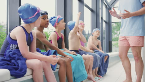 Male-Coach-Giving-Children-In-Swimming-Class-Briefing-As-They-Sit-On-Edge-Of-Indoor-Pool