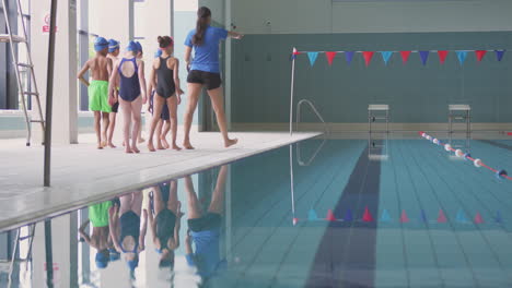 Female-Coach-Walks-Children-In-Swimming-Class-Along-Edge-Of-Indoor-Pool