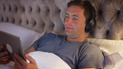 Mann-Mit-Kabellosen-Kopfhörern-Liegt-Im-Bett-Und-Schaut-Vor-Dem-Schlafengehen-Auf-Ein-Digitales-Tablet