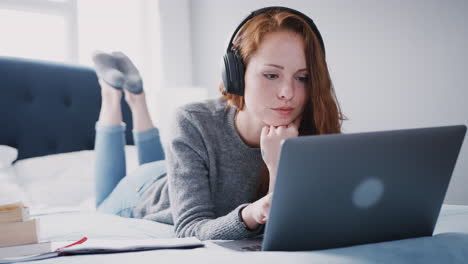 Female-College-Student-Wearing-Headphones-Works-On-Bed-In-Shared-House-With-Laptop-And-Mobile-Phone